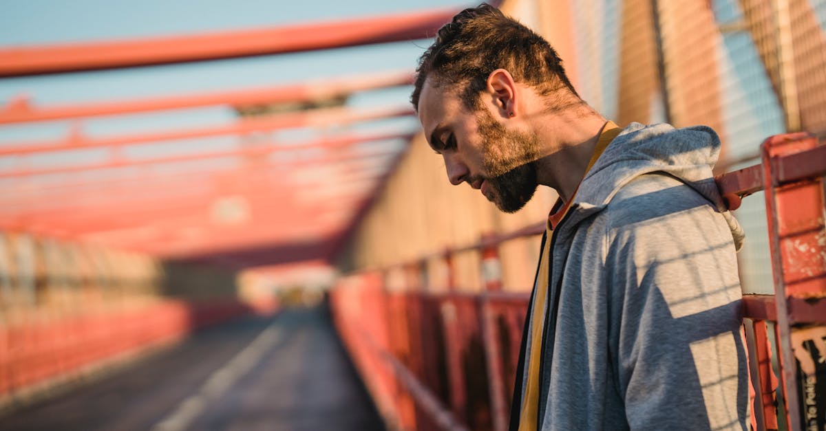 Side view of unshaven millennial guy in hoodie looking down on road of city bridge in sunlight
