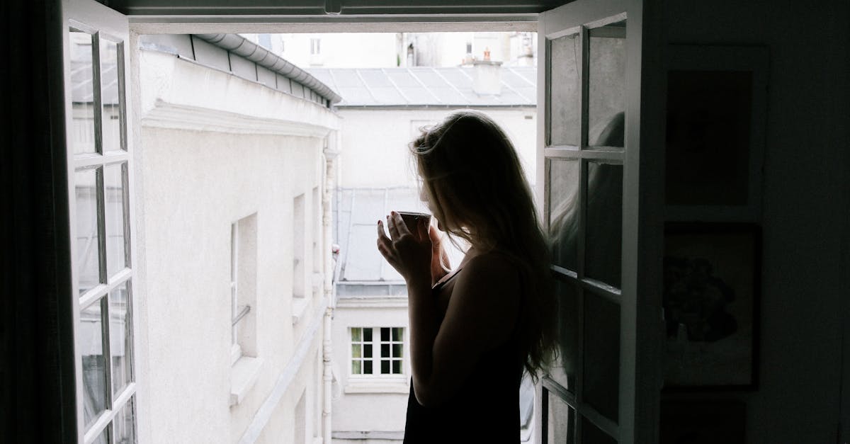 Silhouette of a woman sipping coffee by an open window with a city view.