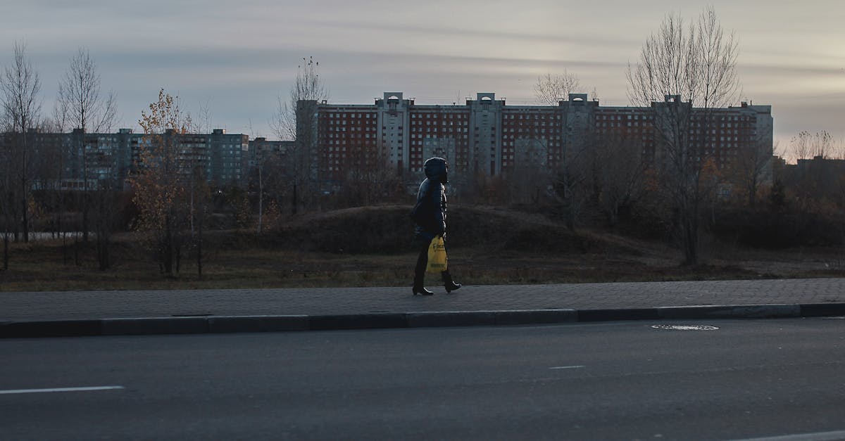 A solitary figure walks on a city sidewalk during dusk, buildings silhouetted in the background.