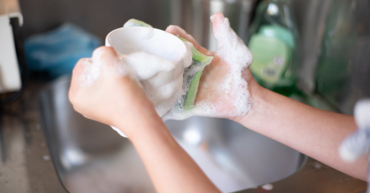 Close-up of hands washing a cup with soap suds in a kitchen sink, representing daily home chores.