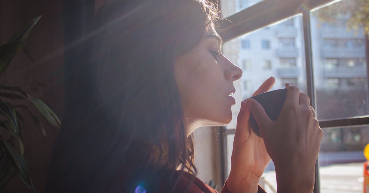 A woman savors a hot drink by a sunlit window in an urban setting.