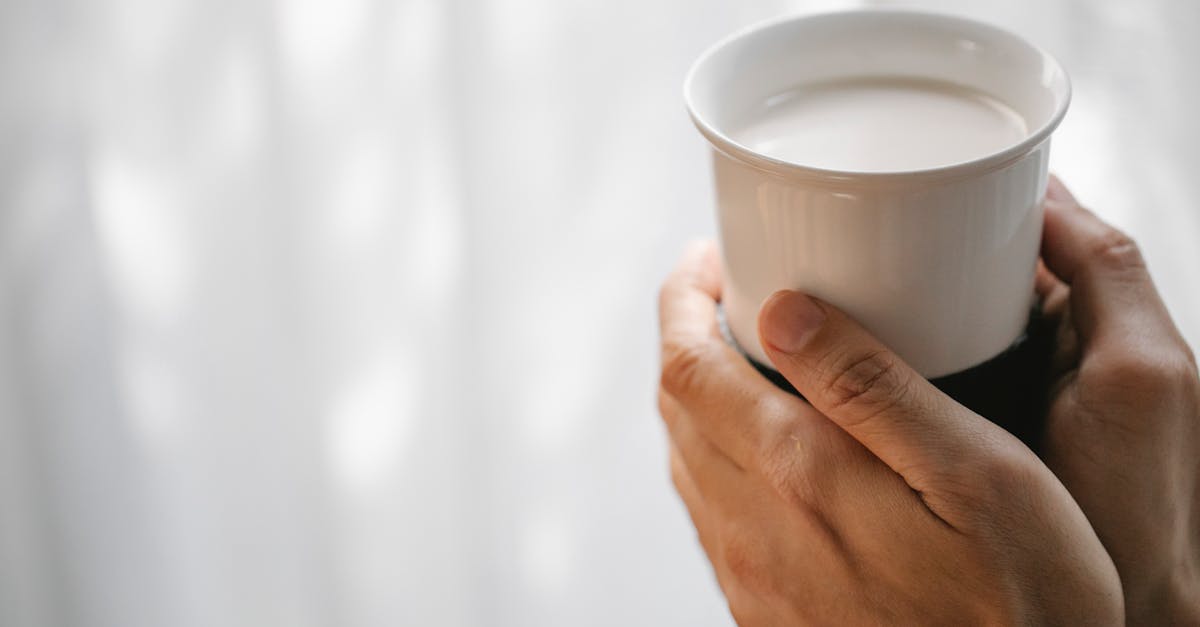 Crop unrecognizable person holding ceramic cup of hot organic milk against blurred light wall