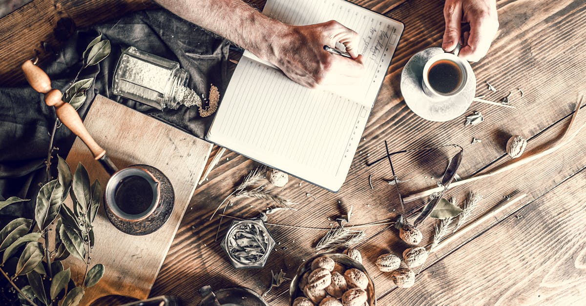 From above senior person taking notes while having coffee at wooden table with scattered walnuts and plants wi