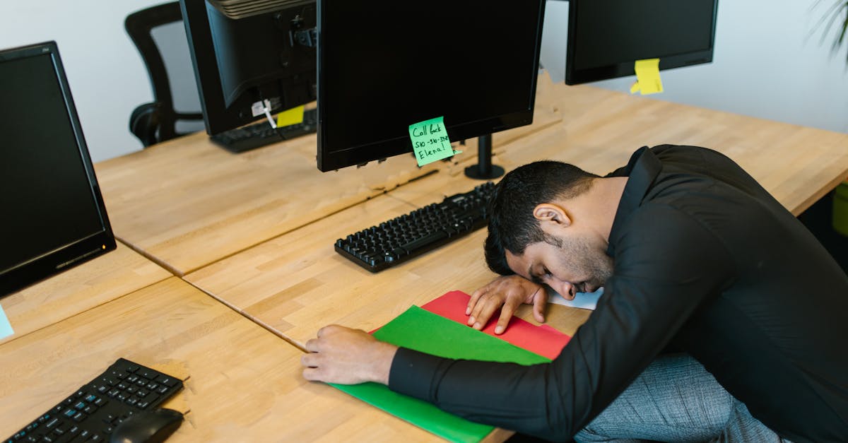 Exhausted office worker falling asleep at desk with monitors and keyboard.
