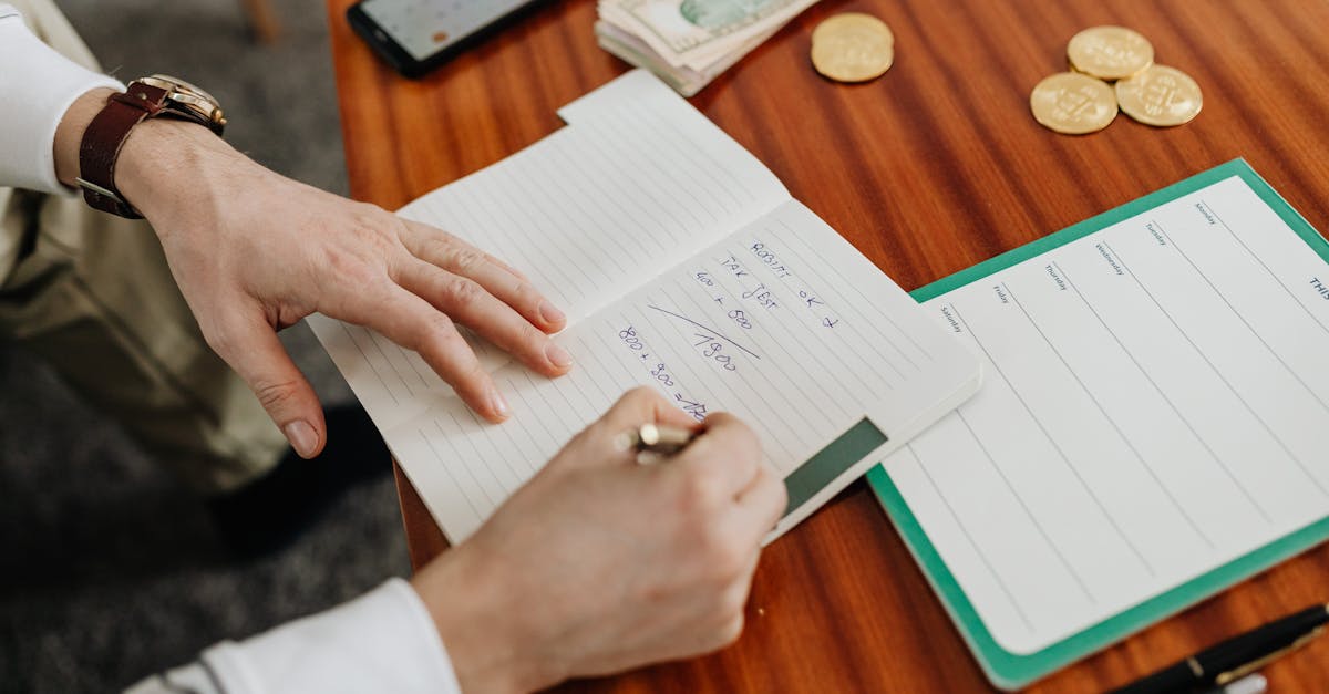 Hands writing financial calculations on notebook with money and coins on table.