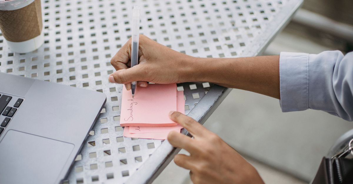 Person writing notes at an outdoor café table with laptop and coffee.