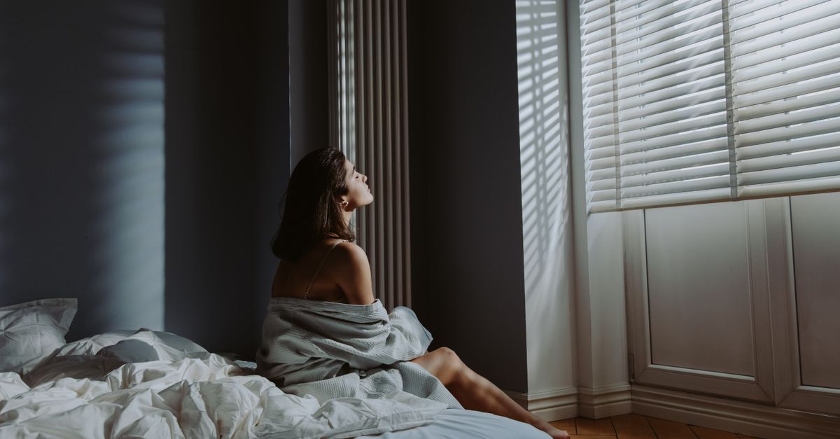 Woman sitting on bed in cozy bedroom, wrapped in a blanket with sunlight streaming through blinds.