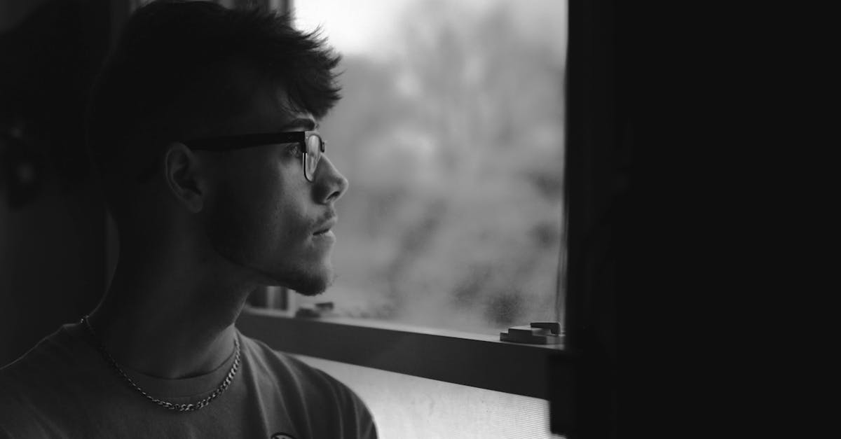 Black and white portrait of a man in eyeglasses looking out a window, feeling pensive.