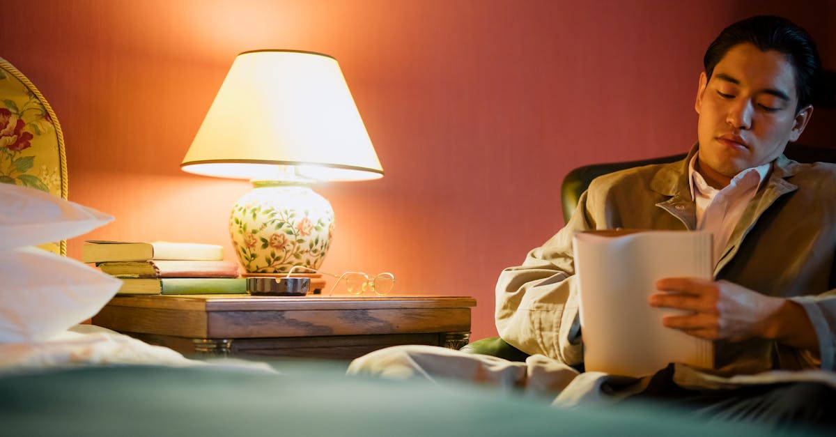An adult man reads in a warmly lit hotel bedroom with a lamp and bedside table.