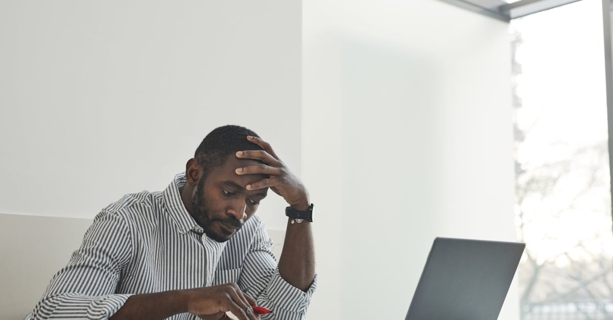 A thoughtful man sitting at a desk with a laptop and papers, contemplating work in a bright, modern office.