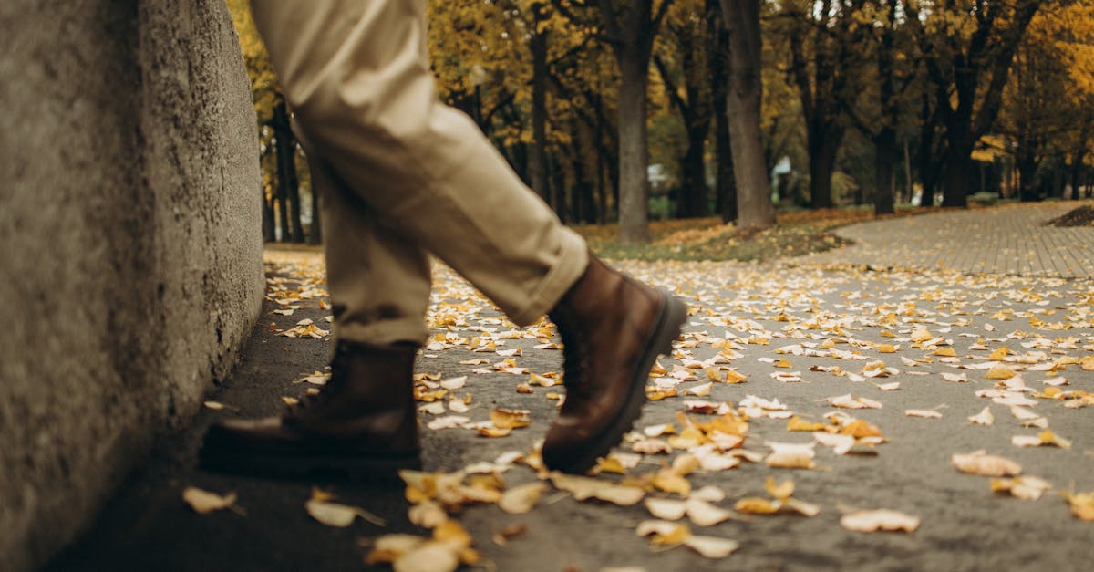 A person in boots walks through a park covered in autumn leaves.