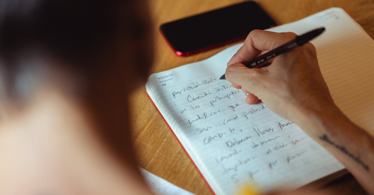 A person writes in a notebook at a wooden desk with a phone nearby.