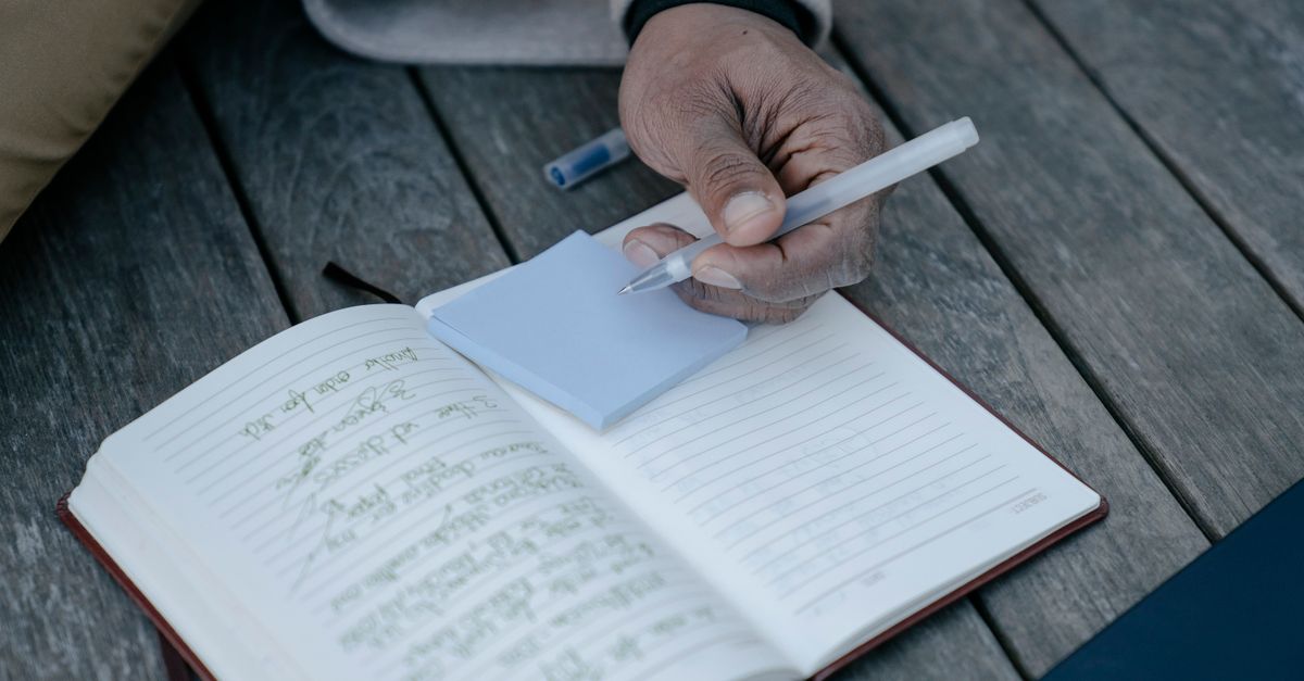 Person writing in a journal, focusing on hand holding pen with notepad on a wooden table.