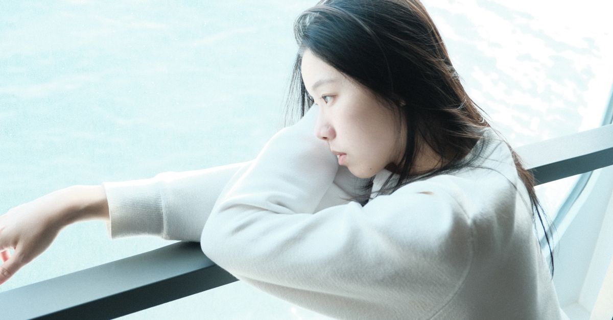 A young woman in a sweatshirt gazes thoughtfully out a window, leaning on a railing.