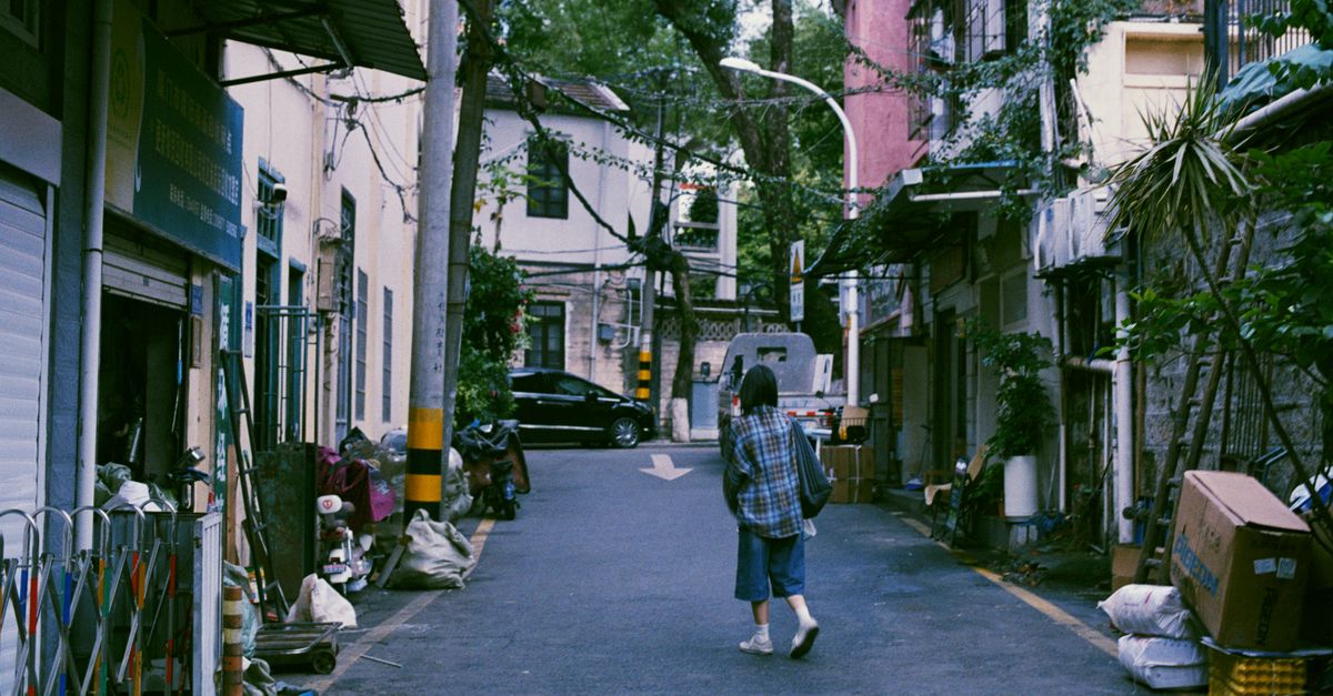 A person in plaid walks through a quiet urban alleyway lined with greenery.