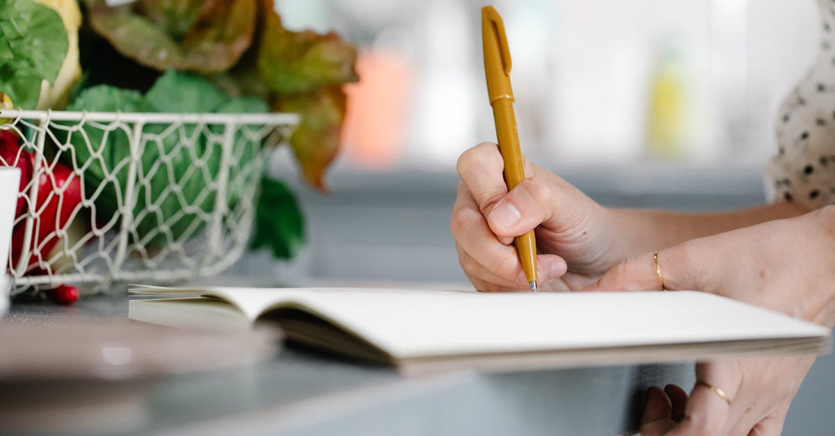 Close-up of a woman writing in a notebook on a kitchen counter beside a basket of fresh vegetables.