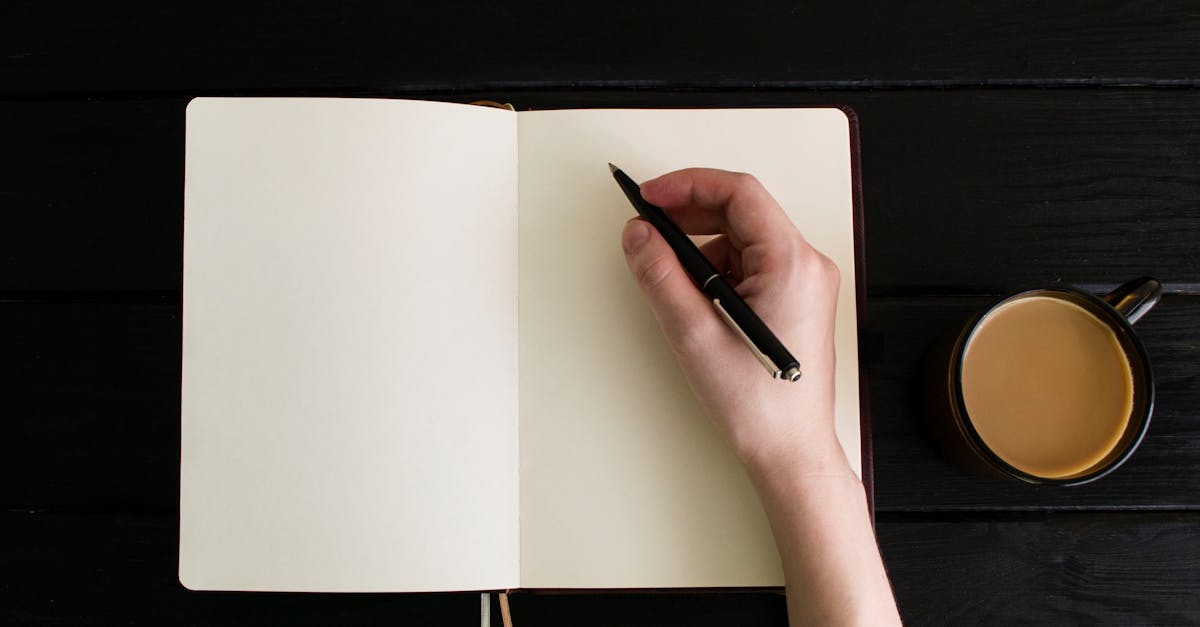 Top view of a hand writing in a blank notebook with a coffee cup on a dark wooden table.