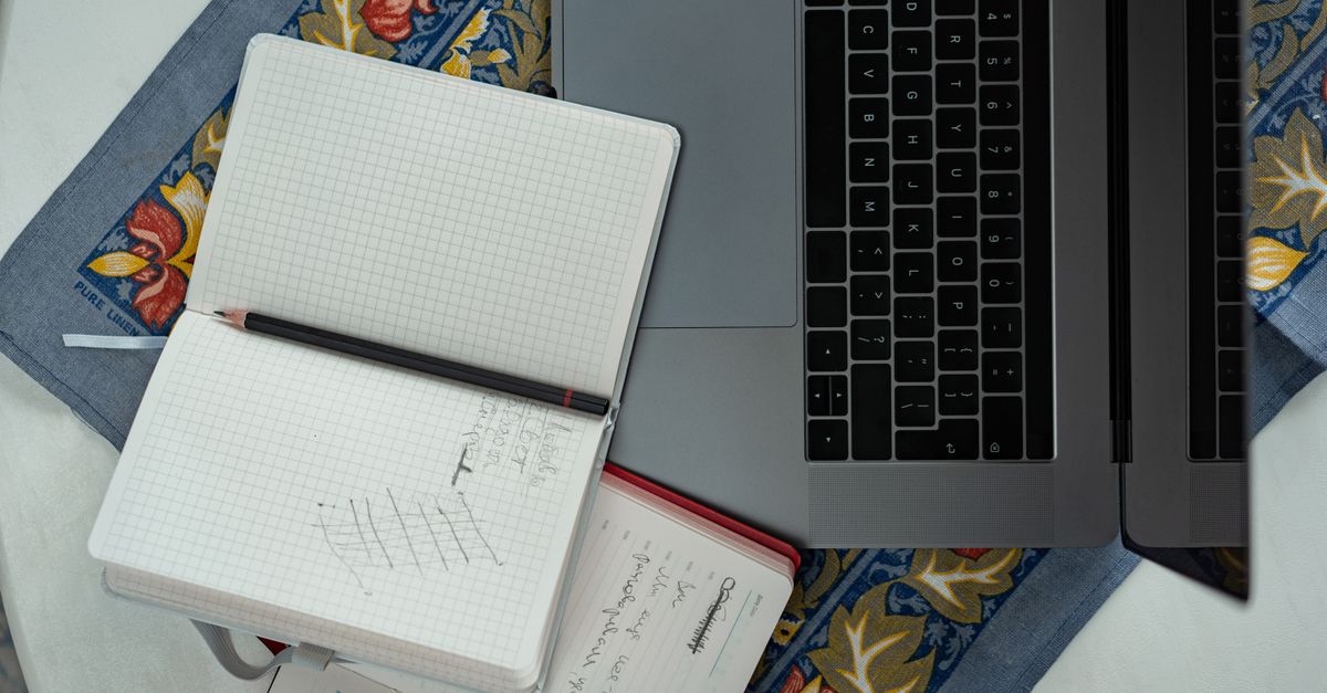 Flat lay of a laptop, notebooks, and a pencil on a floral tablecloth, ideal for productivity themes.