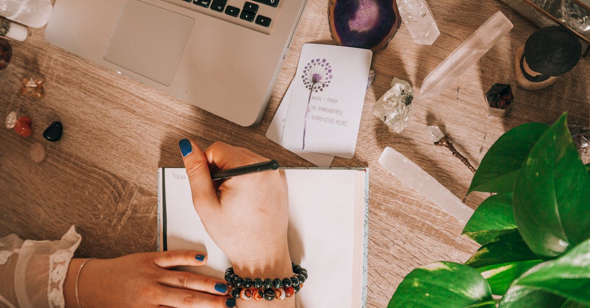 Woman's hand writing in a journal surrounded by crystals, plants, and laptop on a wooden desk.